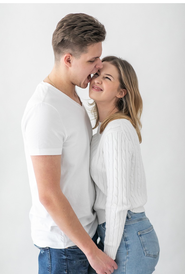 Młoda para w jasnych ubraniach, on próbuje ugryźć jej policzek, ona się uśmiecha. Ujęcie w studio na białym tle, bliskość i emocje.