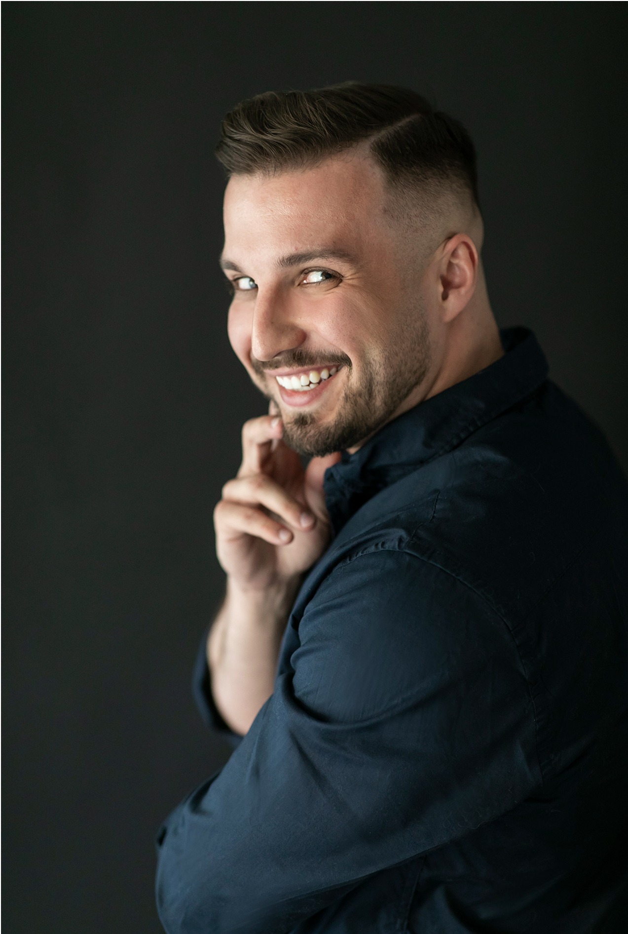 Uśmiechnięty mężczyzna w granatowej koszuli, z modną fryzurą i lekkim zarostem, spogląda przez ramię na czarnym tle.