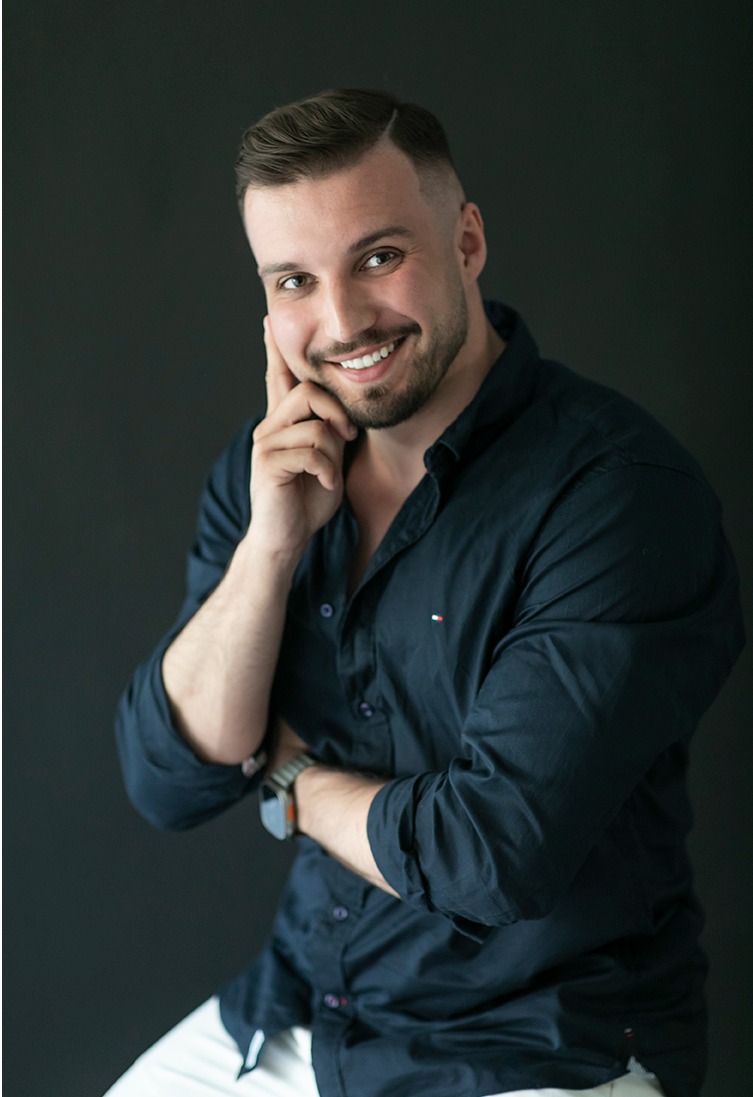 Uśmiechnięty mężczyzna w ciemnej koszuli, z ręką przy twarzy, na ciemnym tle. Portret biznesowy z zegarkiem na ręce i modną fryzurą.