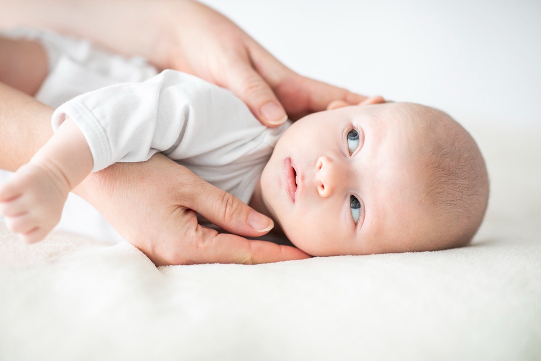 Niemowlę w białym ubranku leży na miękkim, jasnym tle, delikatnie podtrzymywane przez dłonie dorosłego. Uwagę przykuwają jego jasne, niebieskie oczy.