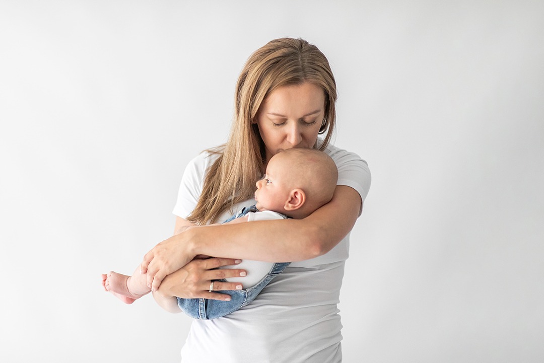 Matka w białej koszulce całuje w głowę niemowlę w dżinsowych ogrodniczkach na tle białej ściany. Delikatne oświetlenie podkreśla więź.