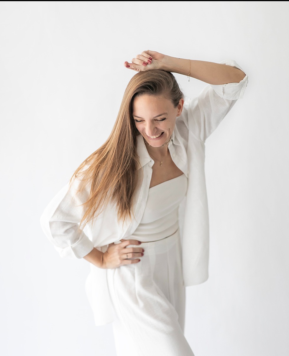 Uśmiechnięta blondynka w białym, luźnym stroju pozuje na jasnym tle w Warszawie. Dłoń na biodrze, druga uniesiona nad głową, subtelny uśmiech, naturalny makijaż.