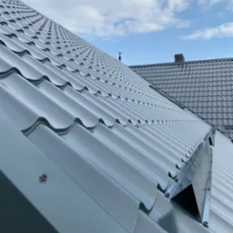 Błyszczący, szary dach z blachy trapezowej w zbliżeniu, widoczne mocowania i fragment rynny. W tle niebo z chmurami i fragment dachu sąsiedniego budynku.