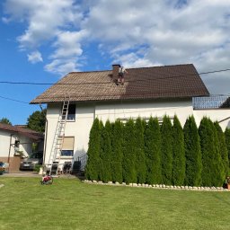 Car Center Waloszczyk Szymon Waloszczyk - Pracownik na brązowym dachu domu podczas pracy. Na trawniku stoi czerwona myjka ciśnieniowa. Biała fasada budynku i rząd zielonych tuj w tle.