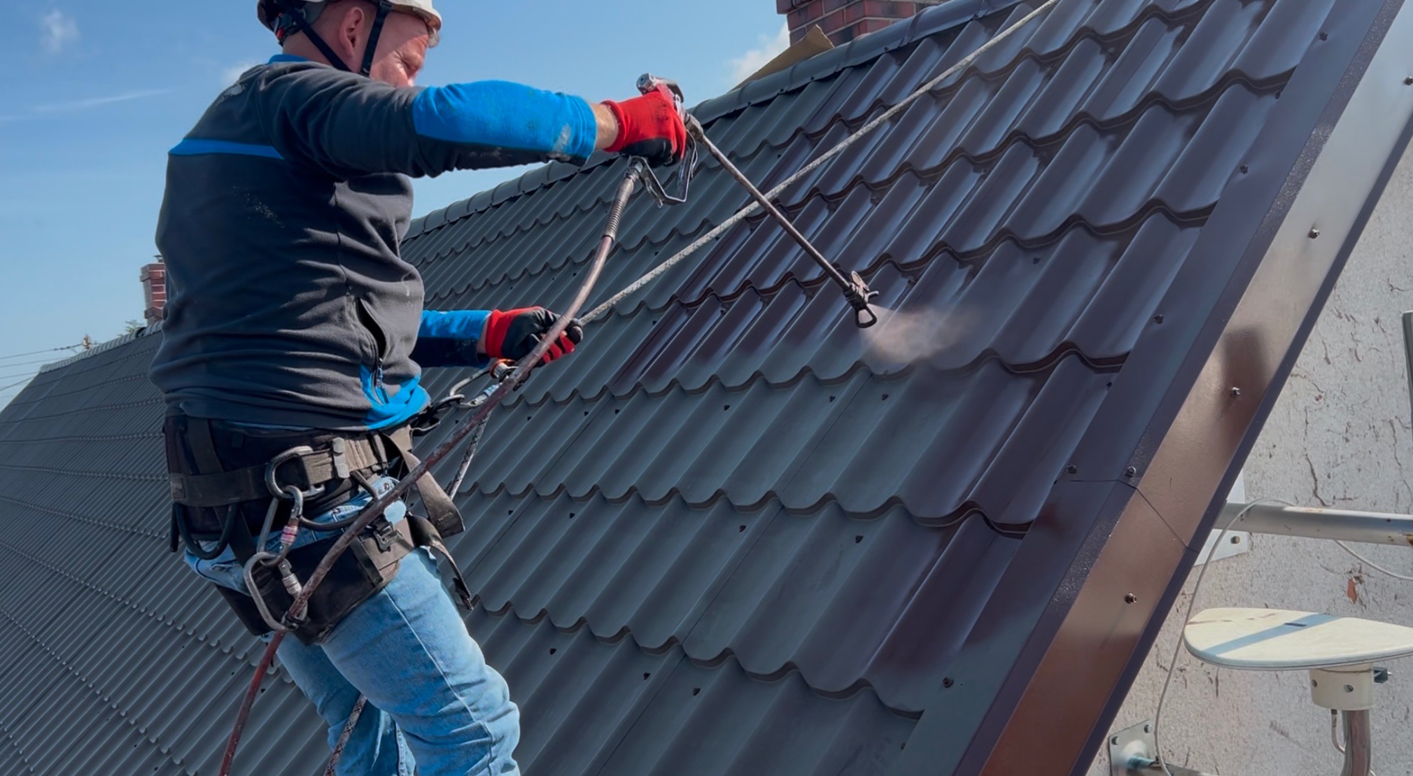 Pracownik w uprzęży maluje brązowy dach z blachodachówki za pomocą pistoletu natryskowego. Widoczne zabezpieczenia i fragment komina.