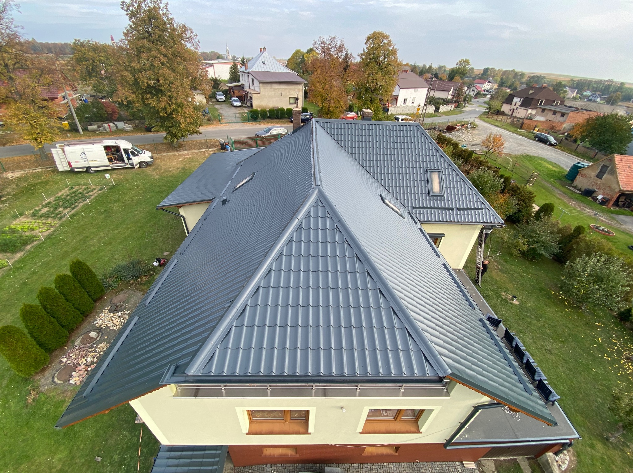 Szary dach z blachodachówki, widok z góry na dom z ogrodem. Na trawniku zaparkowany biały bus. Widoczne elementy architektury podmiejskiej w tle.