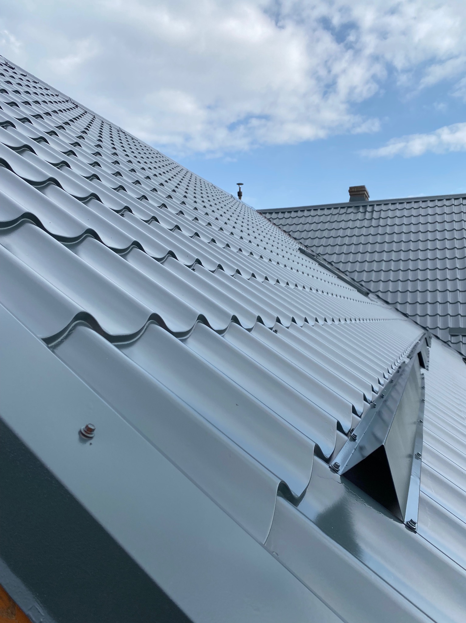 Błyszczący, szary dach z blachy trapezowej w zbliżeniu, widoczne mocowania i fragment rynny. W tle niebo z chmurami i fragment dachu sąsiedniego budynku.