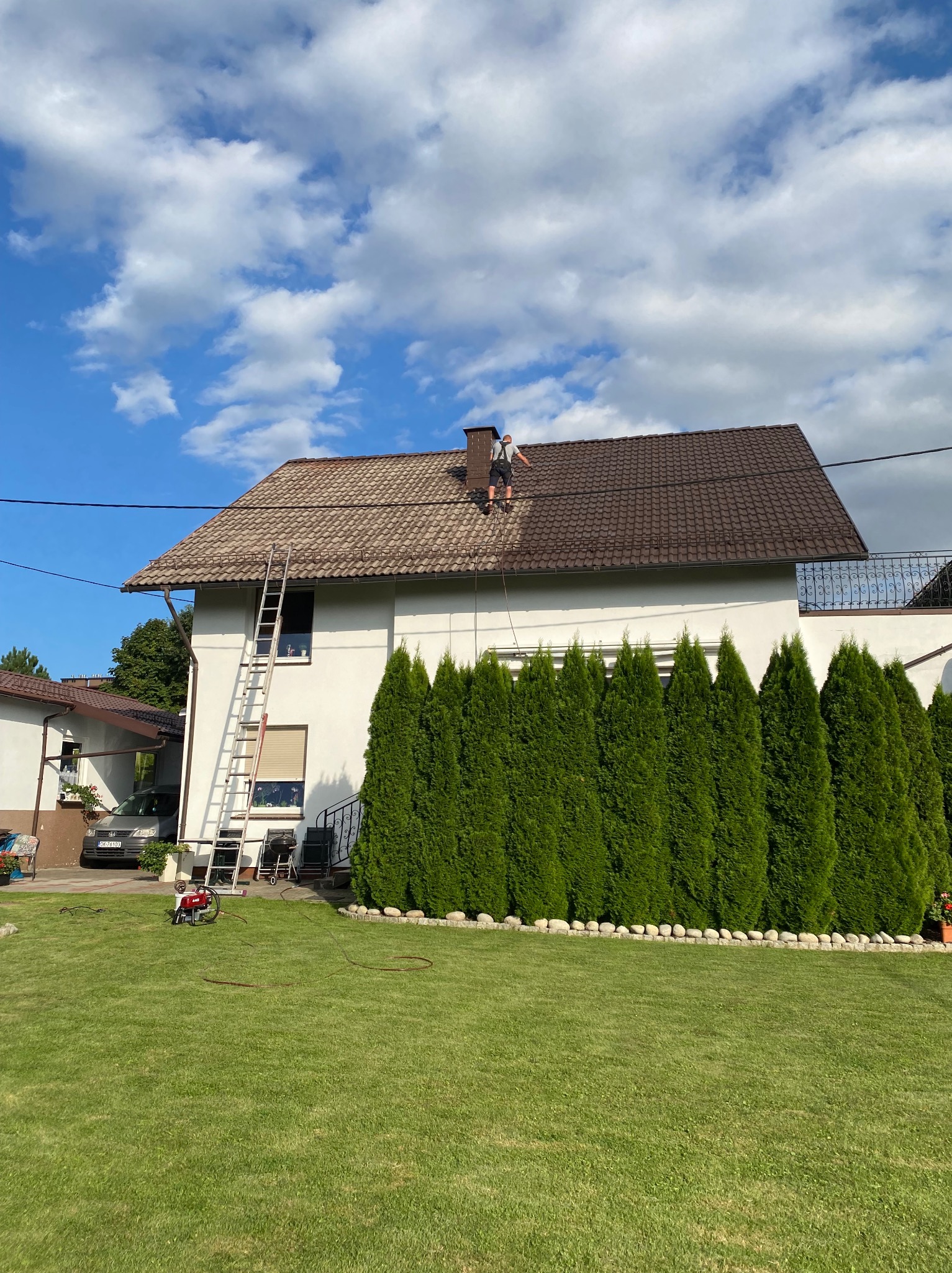 Pracownik na brązowym dachu domu podczas pracy. Na trawniku stoi czerwona myjka ciśnieniowa. Biała fasada budynku i rząd zielonych tuj w tle.