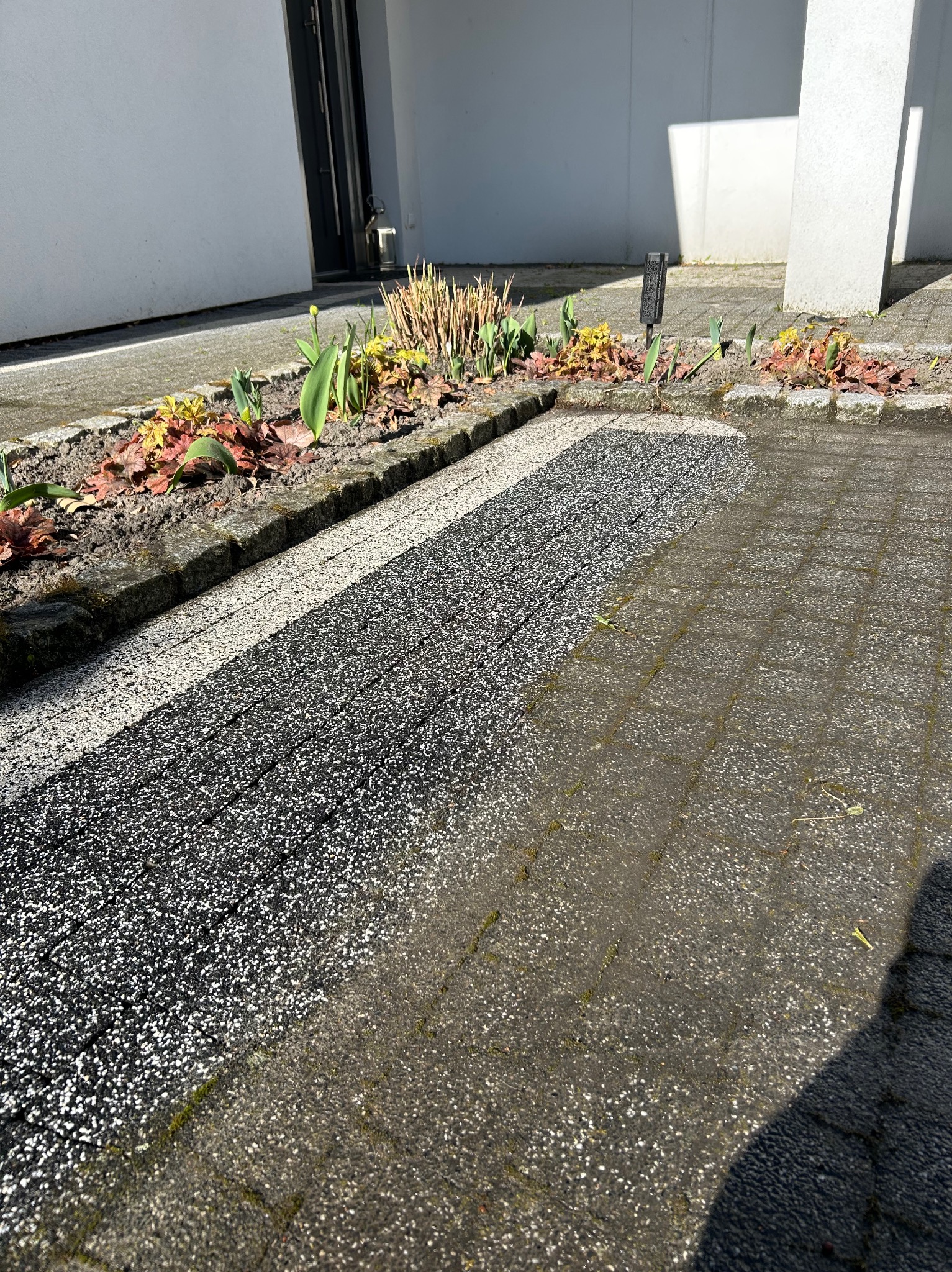 Częściowo wyczyszczona kostka brukowa przed budynkiem, z widocznym fragmentem rabaty kwiatowej i elementami elewacji w tle, w słoneczny dzień.