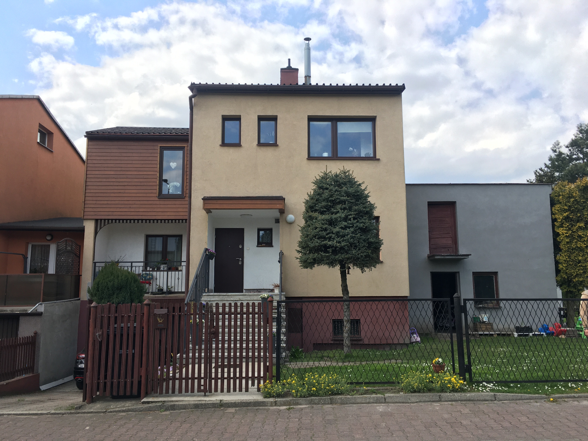 Dwupiętrowy dom z beżową elewacją i brązowymi akcentami, otoczony ogrodzeniem. W oknie widoczna dekoracja w kształcie serca. Na pierwszym planie drzewko.