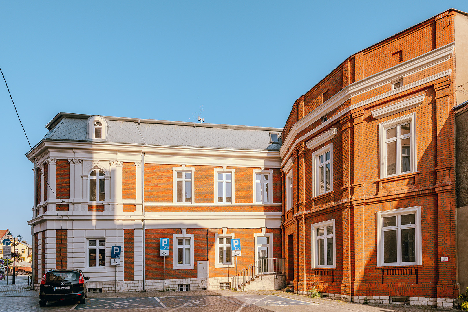 Odrestaurowany budynek z czerwonej cegły z białymi detalami architektonicznymi i szarym dachem, widoczny z parkingu z zaparkowanym autem.