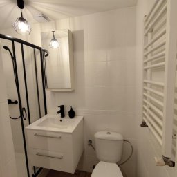 Techbruk - Biała łazienka z kabiną prysznicową, umywalką z szafką, toaletą i grzejnikiem drabinkowym. Minimalistyczny design, czarne akcenty, drewnopodobna podłoga.