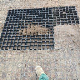 Techbruk - Układanie plastikowej kratki trawnikowej na ziemi, częściowo wypełnionej ziemią, widoczna noga w roboczym ubraniu. Montaż nawierzchni przepuszczalnej.