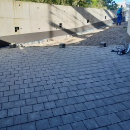 Techbruk - Ułożona, szara kostka brukowa na zewnątrz budynku z betonową ścianą w tle. Widoczne przygotowane podłoże i narzędzia. Prace brukarskie w toku.