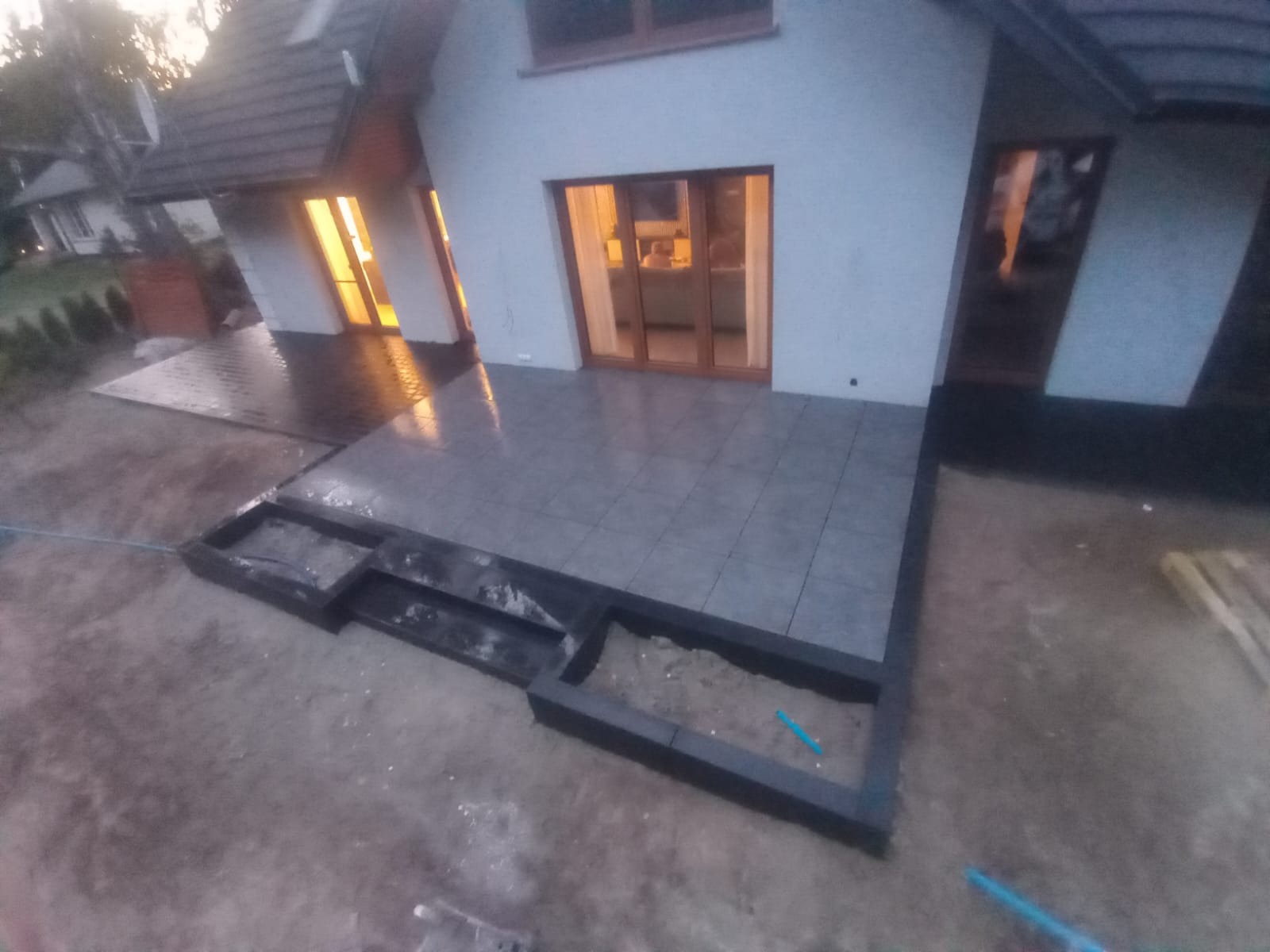 Ułożona szara płytka na tarasie domu jednorodzinnego z czarnymi obrzeżami rabatki. Widoczne otwarte drzwi balkonowe i okna z ciepłym światłem wewnątrz.