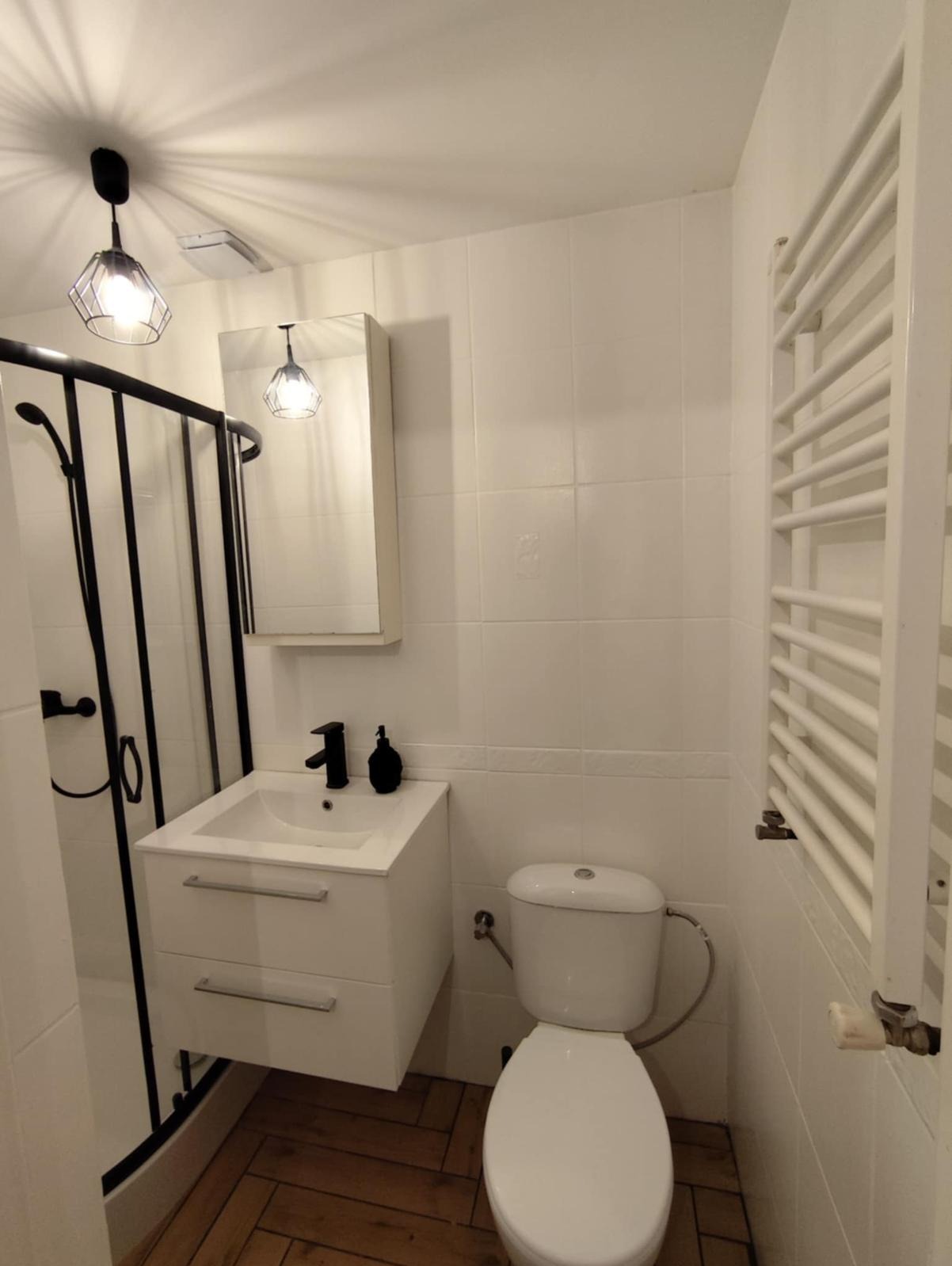 Biała łazienka z kabiną prysznicową, umywalką z szafką, toaletą i grzejnikiem drabinkowym. Minimalistyczny design, czarne akcenty, drewnopodobna podłoga.