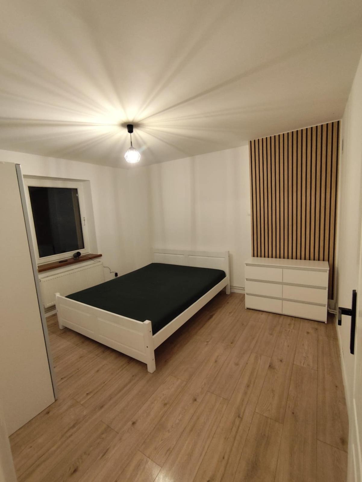 Pokój po wykończeniu: białe meble (łóżko, komoda), drewniane panele na ścianie, podłoga z paneli, nowoczesna lampa sufitowa. Minimalistyczny design.