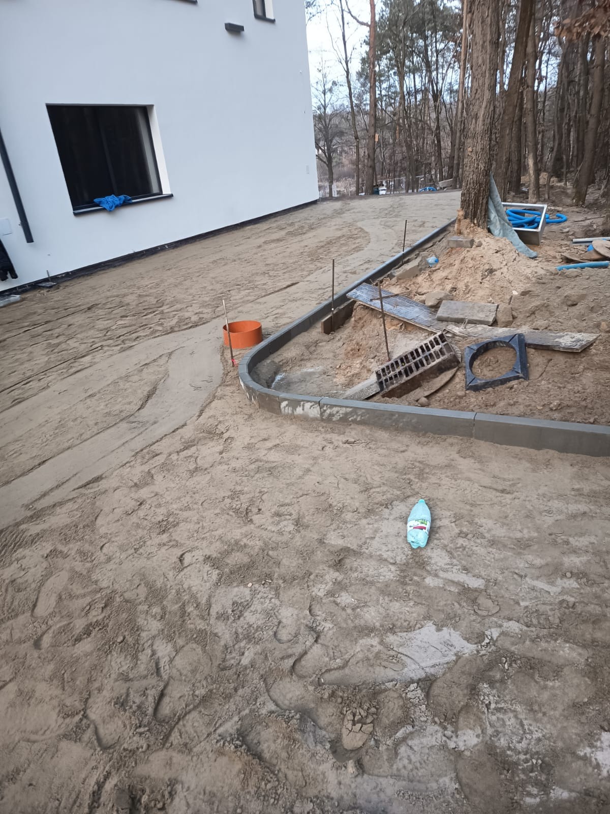 Układanie obrzeży chodnika z kostki betonowej przy nowym domu. Widoczne przygotowane podłoże, elementy kanalizacji deszczowej i butelka wody. Prace ziemne w toku.