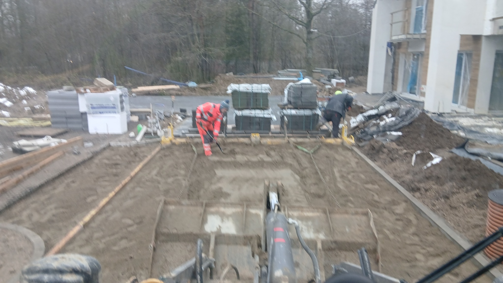 Układanie kostki brukowej na posesji. Dwóch pracowników w odzieży roboczej przygotowuje podłoże pod kostkę. Widok z perspektywy operatora maszyny budowlanej.