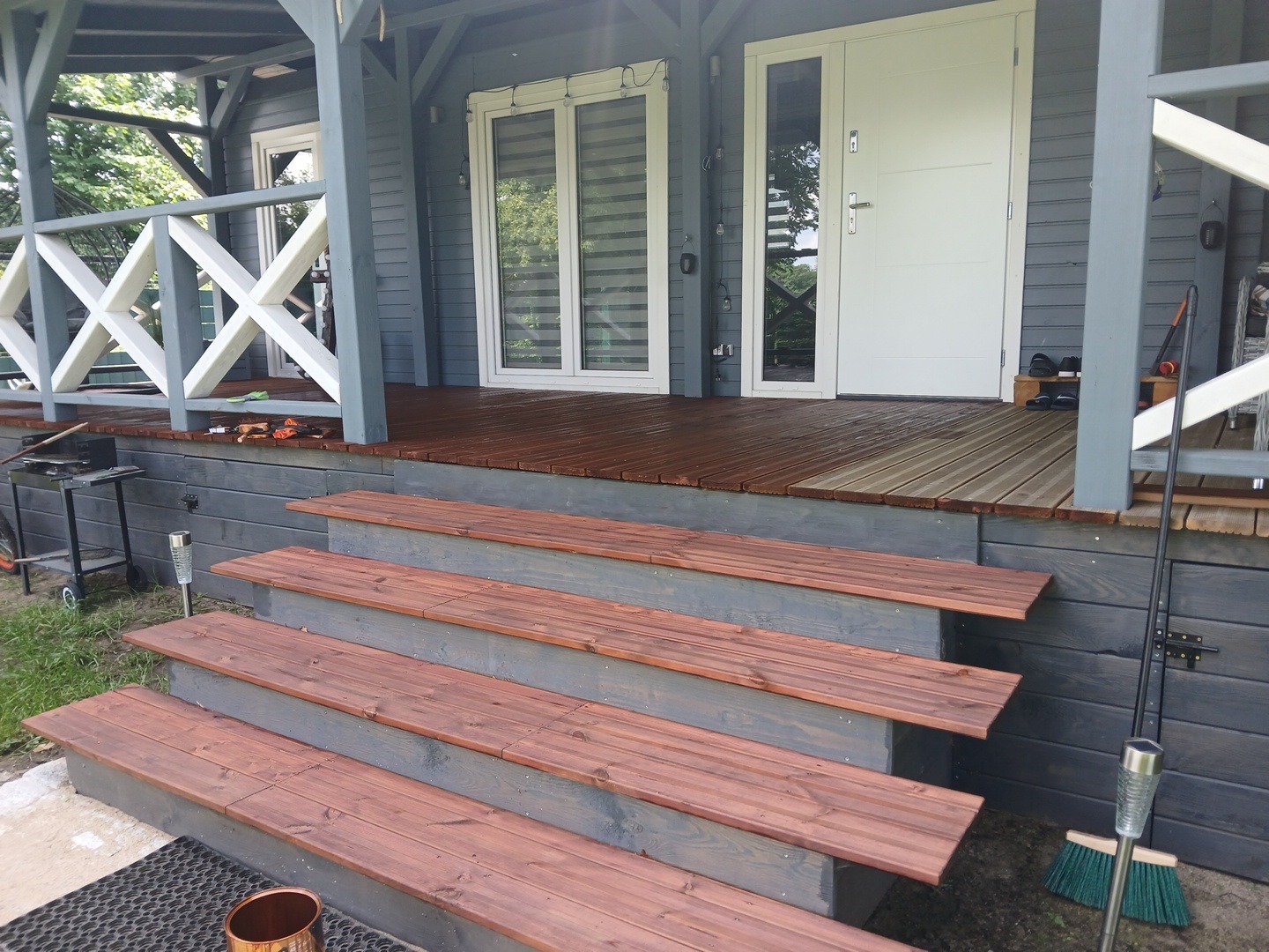 Drewniany taras z pomalowanymi schodami wejściowymi do domu w odcieniach szarości i brązu, widok z dołu, barierka z białymi krzyżami, drzwi wejściowe.