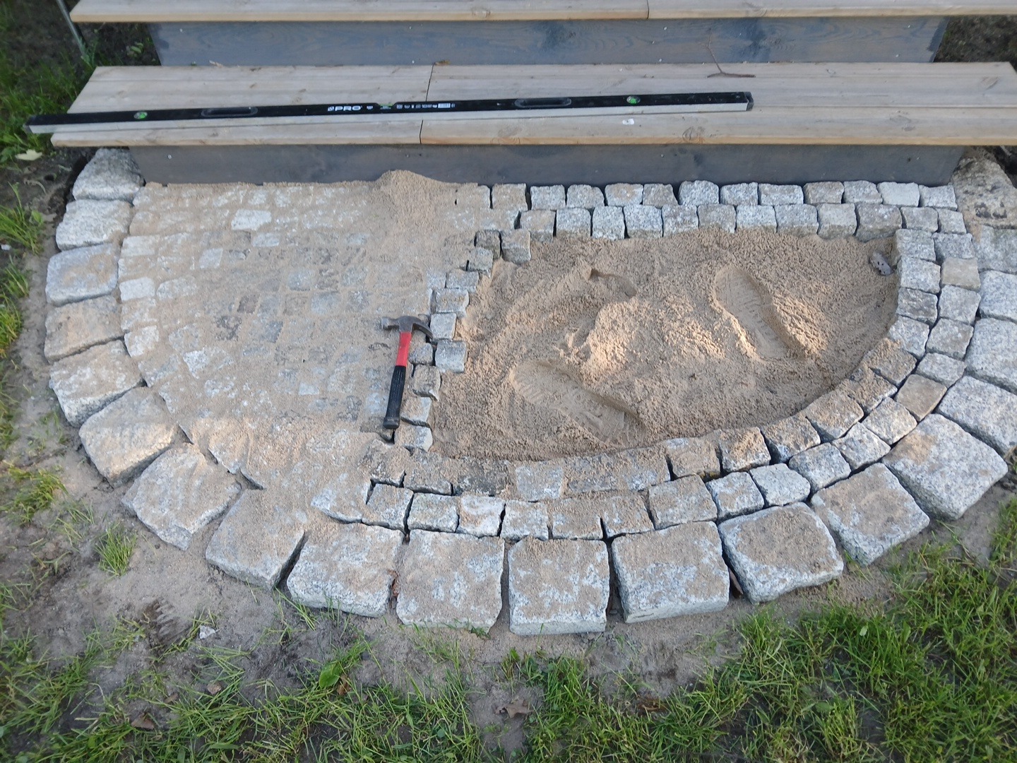 Półokrągła nawierzchnia z kostki granitowej w trakcie układania, widoczny piasek, poziomica i młotek. Etap prac przy schodach zewnętrznych.