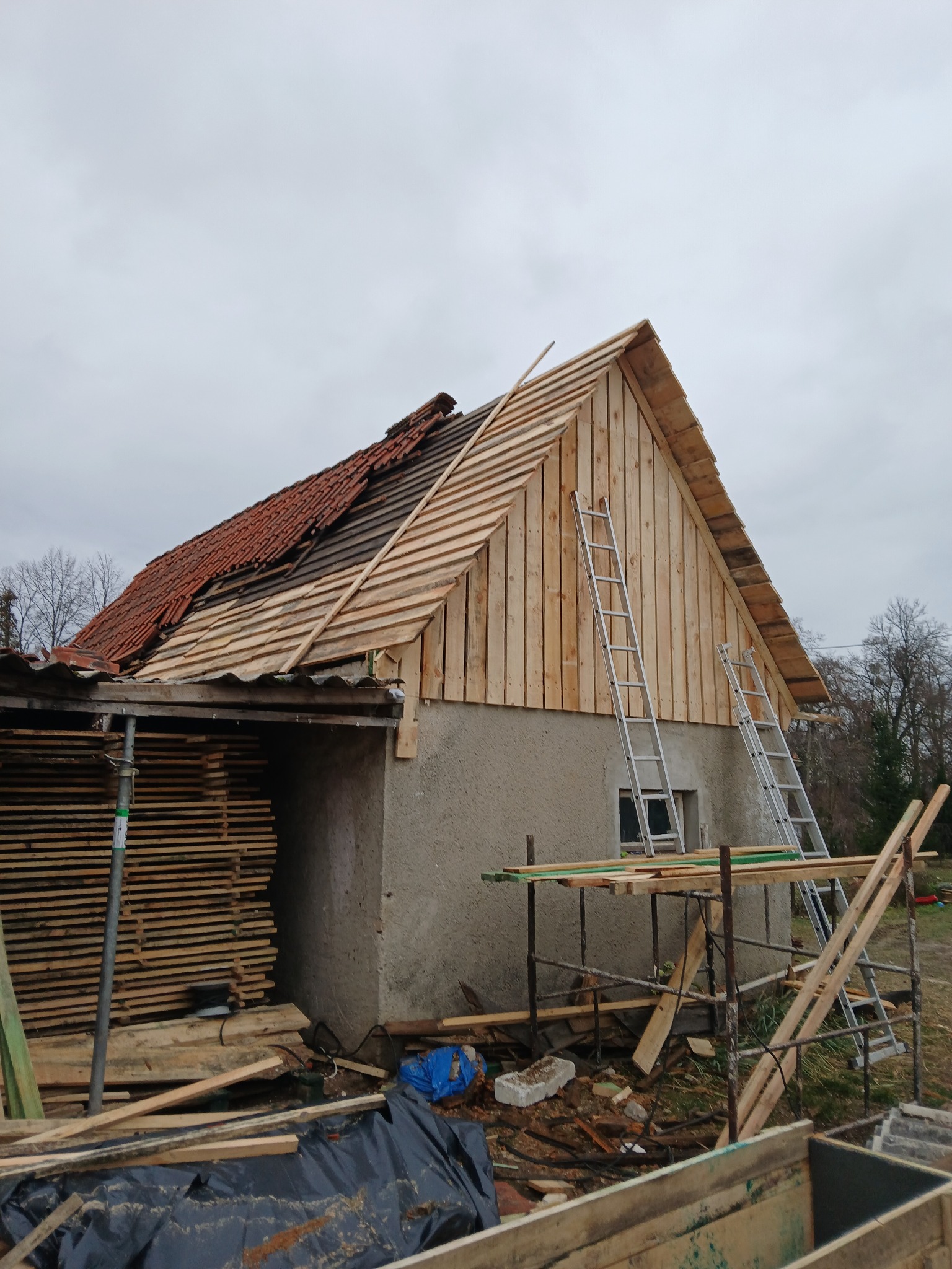 Remont dachu domu jednorodzinnego w trakcie wymiany pokrycia. Widoczne rusztowania, drabiny i deski. Część dachu z nowym drewnianym poszyciem, część z czerwoną dachówką.