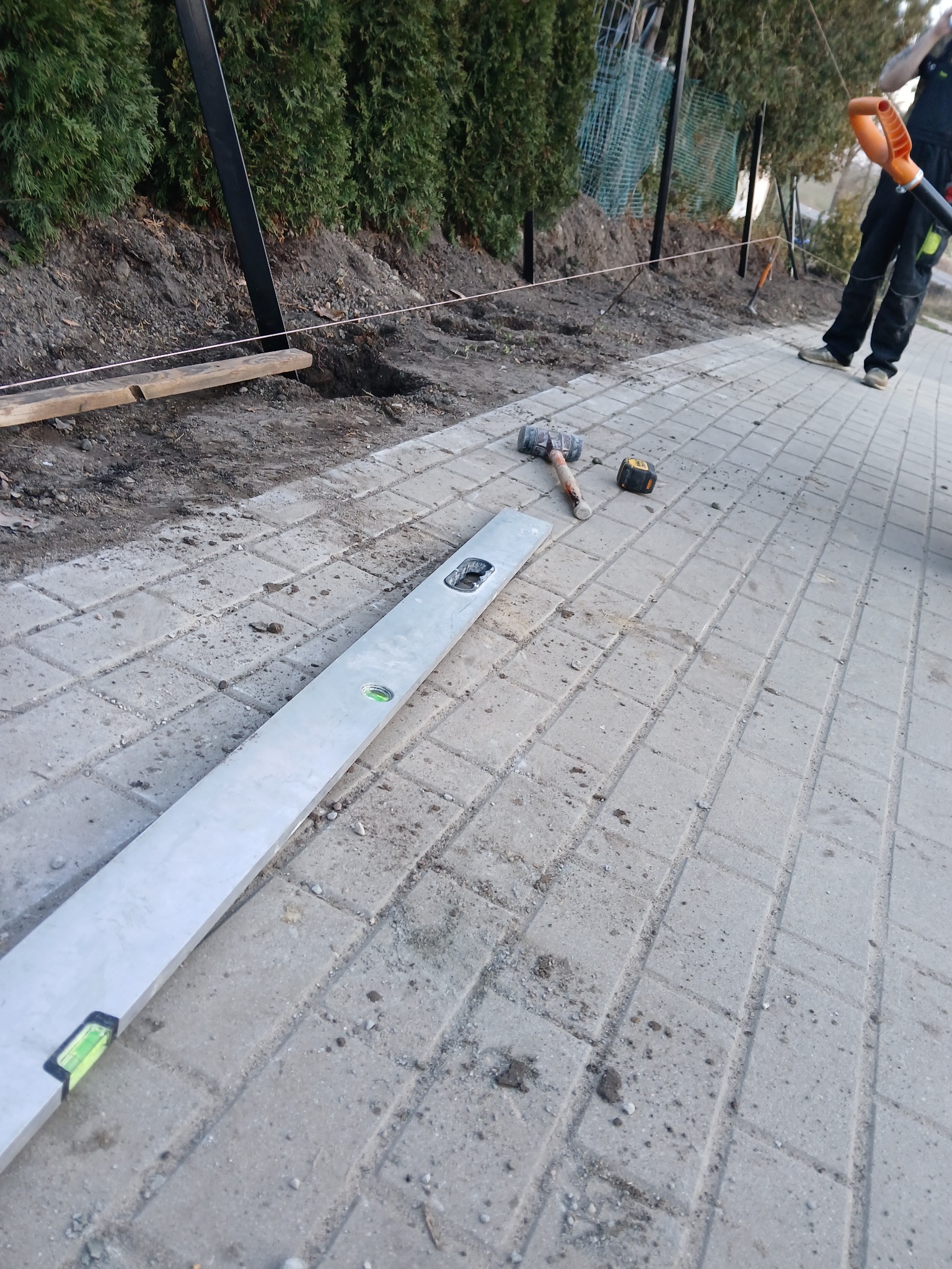 Układanie kostki brukowej: poziomica, młotek i miarka na szarym bruku. W tle ziemia, ogrodzenie i pracownik z narzędziem.