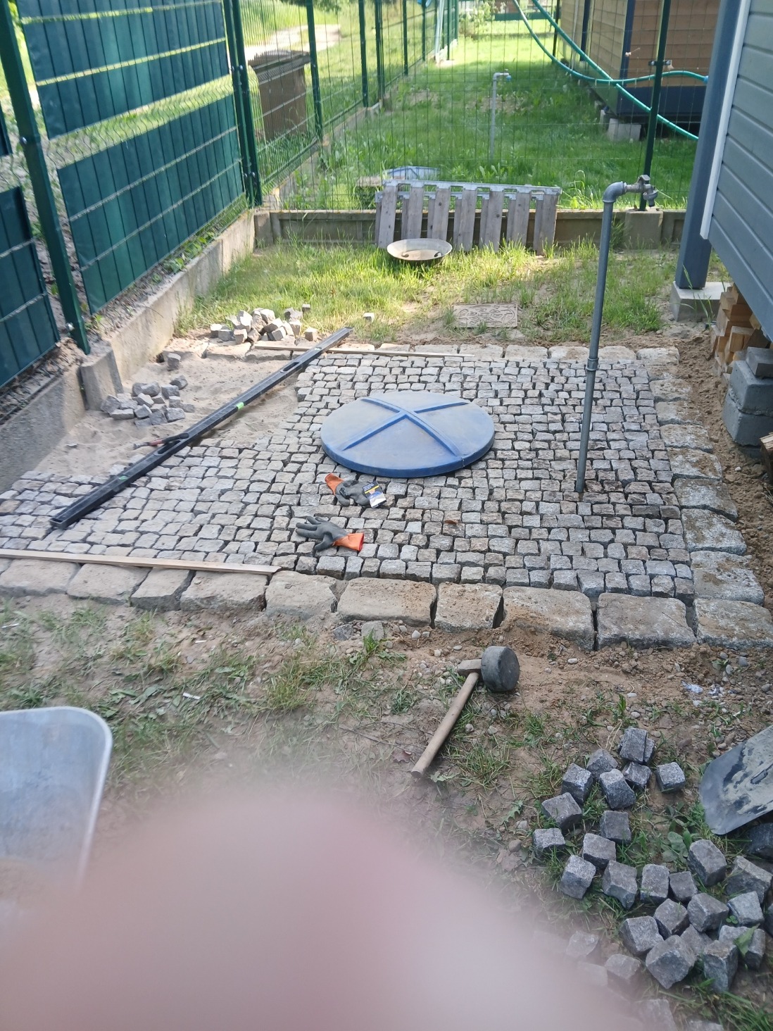 Układanie kostki brukowej wokół studzienki na trawniku, widoczne narzędzia i materiały budowlane. Etap prac brukarskich w ogrodzie.