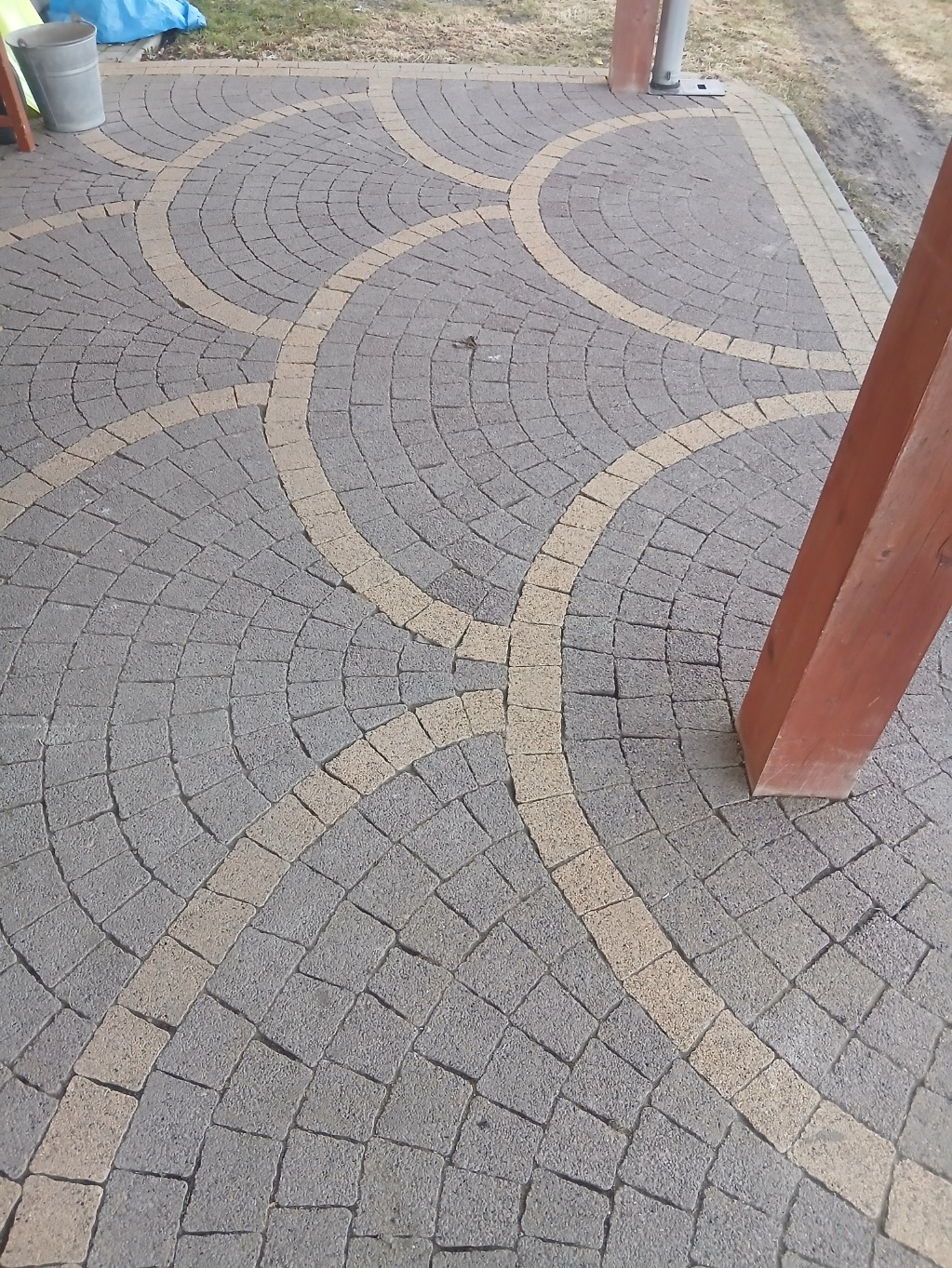 Ułożona kostka brukowa w odcieniach szarości i beżu, tworząca wzór łuków. Fragment drewnianej konstrukcji w prawym dolnym rogu. Widok z góry.