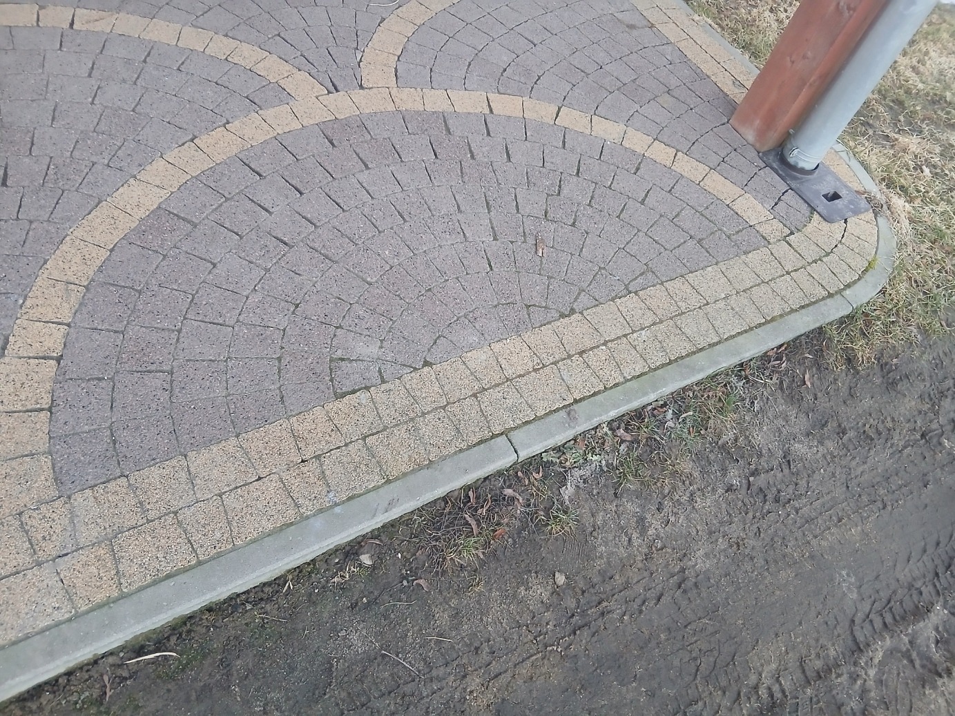 Ułożona kostka brukowa w kształcie półkola z obrzeżem, dwukolorowa, szaro-beżowa, przy słupku i ziemi z resztkami trawy. Widok z góry.