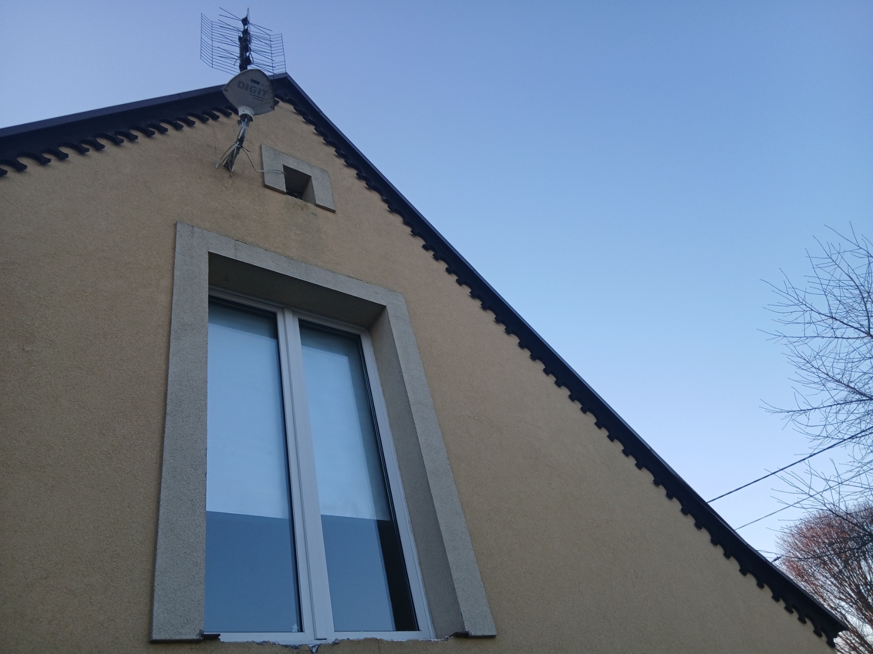 Elewacja domu z oknem i anteną satelitarną na szczycie, widok z dołu, błękitne niebo w tle. Charakterystyczne wykończenie dachu.