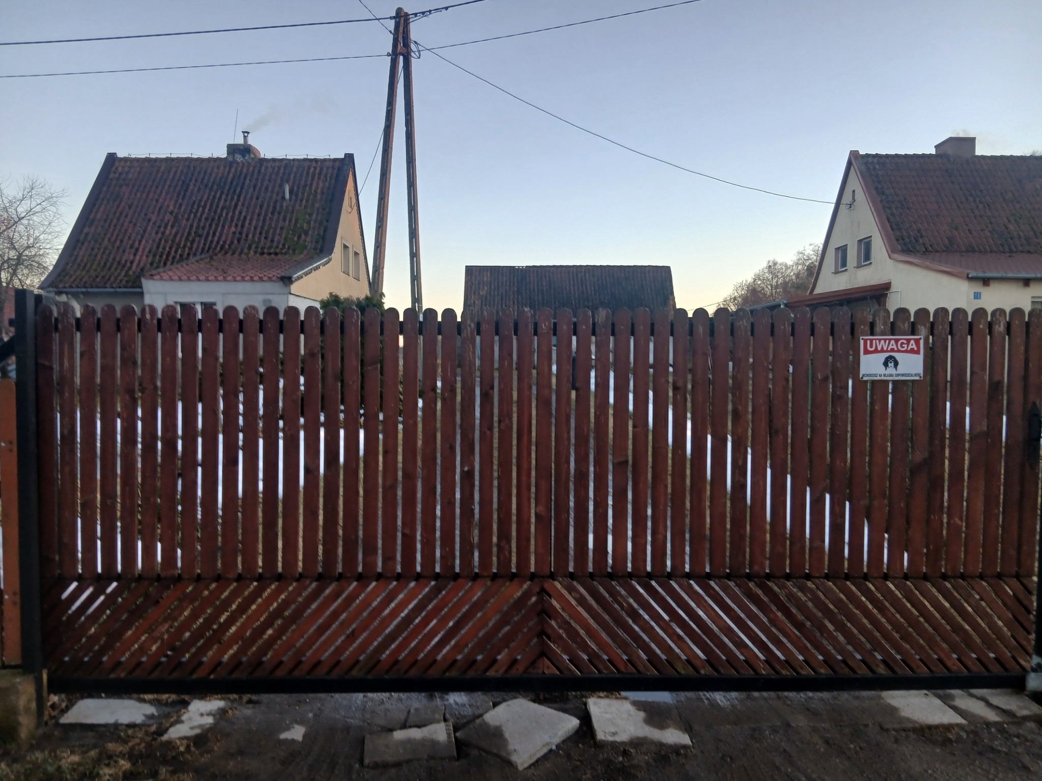 Drewniana brama wjazdowa z pionowymi deskami i ukośnym wzorem u dołu, na tle domów z czerwonymi dachami i słupa energetycznego. Widoczny znak ostrzegawczy.