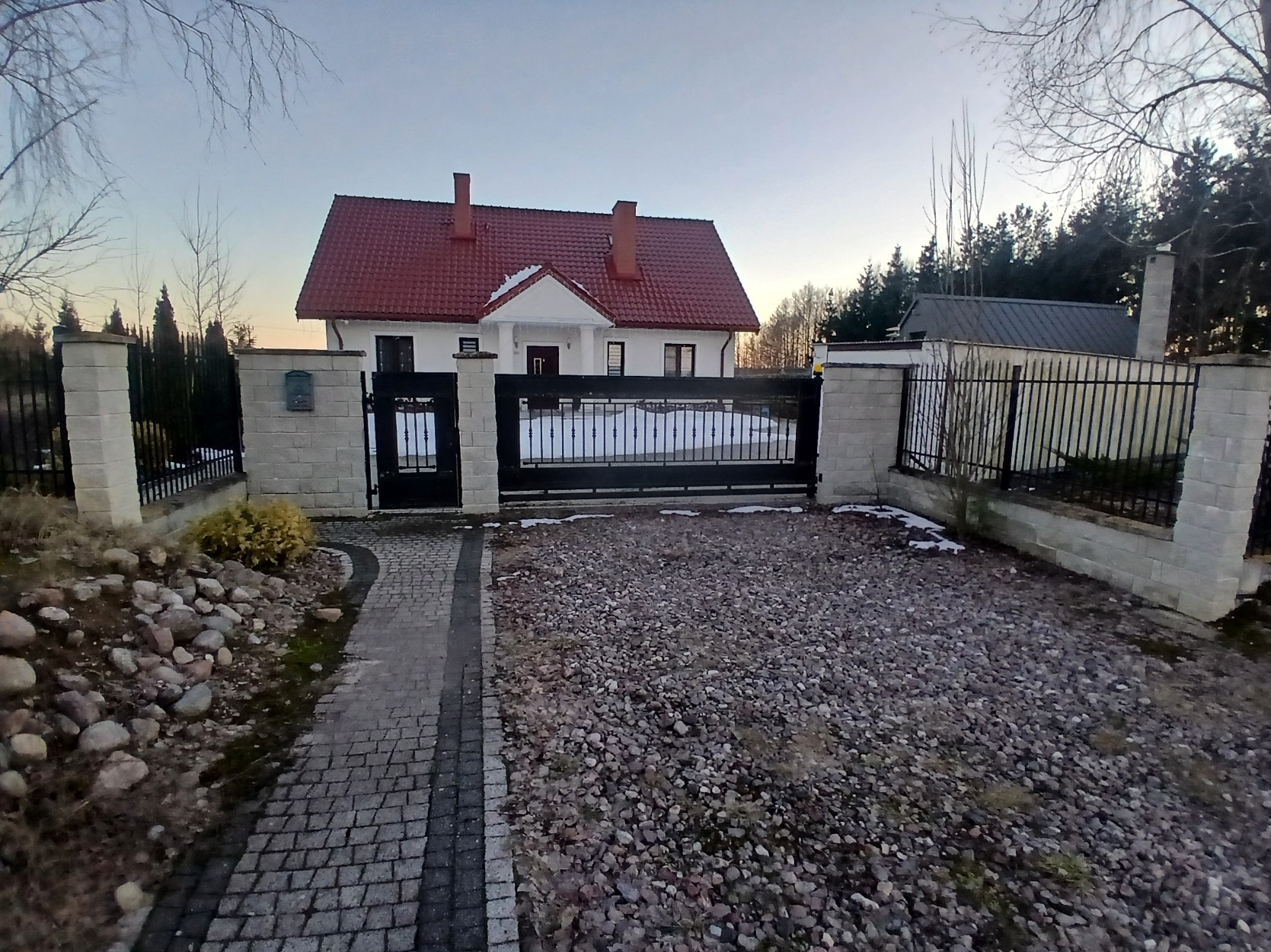 Brama wjazdowa z furtką, murowane słupki z szarej kostki, podjazd wysypany kamieniem, w tle biały dom z czerwoną dachówką i fragment ogrodu.