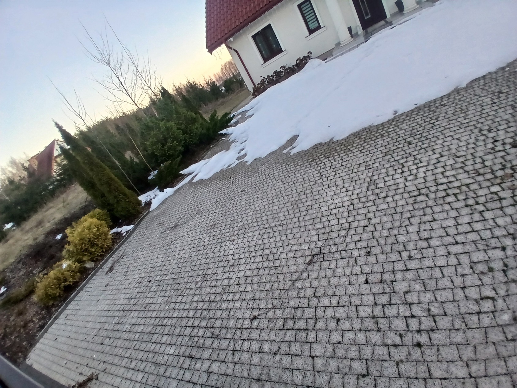 Ułożona kostka brukowa na podjeździe domu jednorodzinnego w zimowej scenerii. Widoczne krzewy i częściowo ośnieżony teren. Ujęcie z niskiej perspektywy.