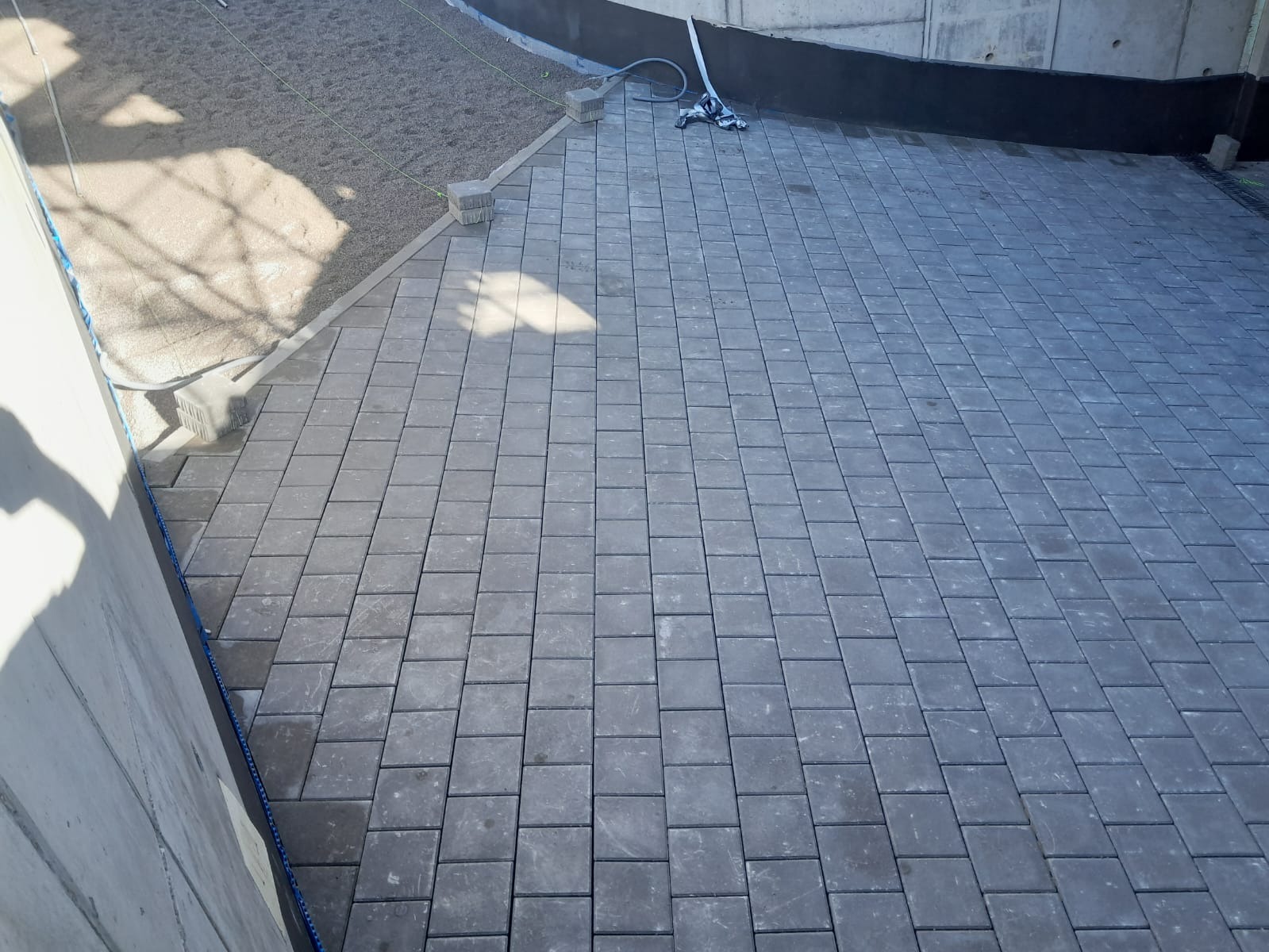 Ułożona szara kostka brukowa na podwórku, widok z góry. Częściowo widoczny teren przygotowany pod dalsze prace brukarskie. W tle betonowy mur.