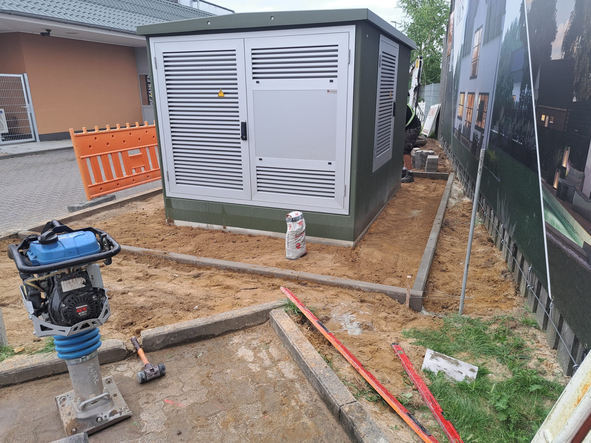 Przygotowanie terenu pod stację transformatorową: ubijarka, obrzeża betonowe, piasek, poziomica. Widoczny fragment budynku i ogrodzenie z reklamą.