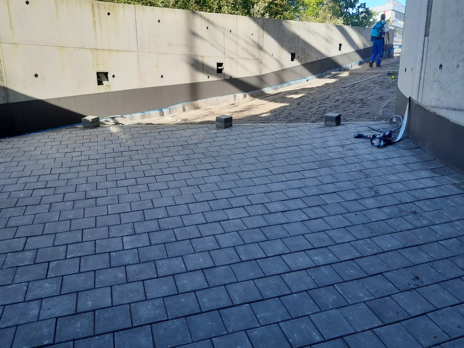 Ułożona, szara kostka brukowa na zewnątrz budynku z betonową ścianą w tle. Widoczne przygotowane podłoże i narzędzia. Prace brukarskie w toku.