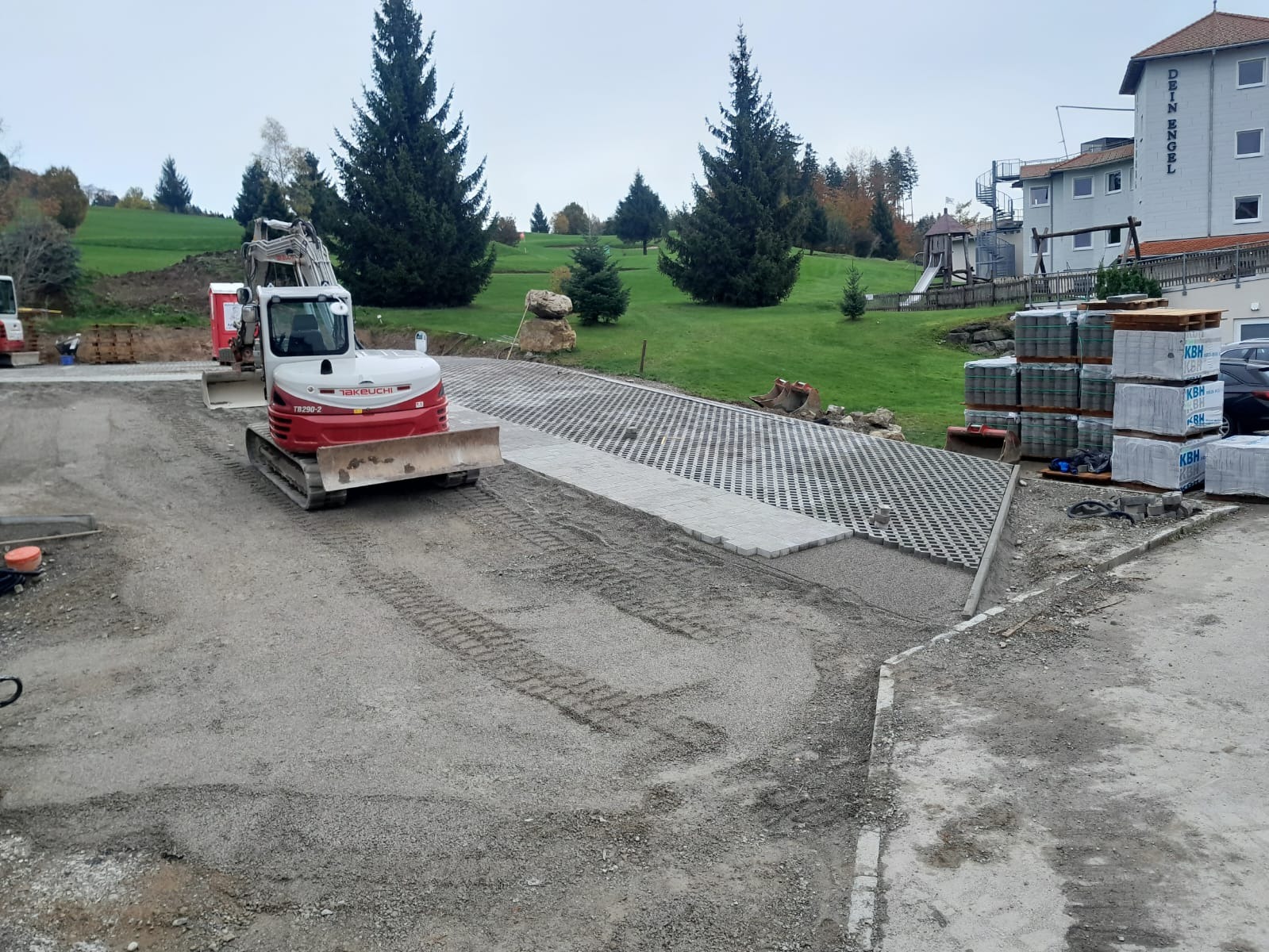 Układanie ażurowych płyt betonowych na podjeździe z użyciem minikoparki Takeuchi TB290-2. W tle budynek z napisem 'DEIN ENGEL' i zielony teren.