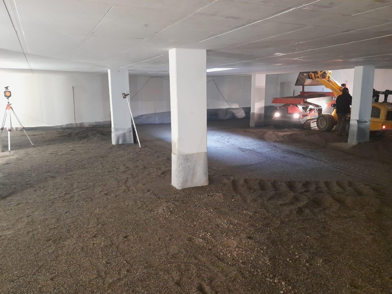 Wyrównywanie podłoża w hali z pomocą miniładowarki i niwelatora laserowego. Przygotowanie do wylewki. Widoczne ślady opon na piasku i kolumny podtrzymujące strop.