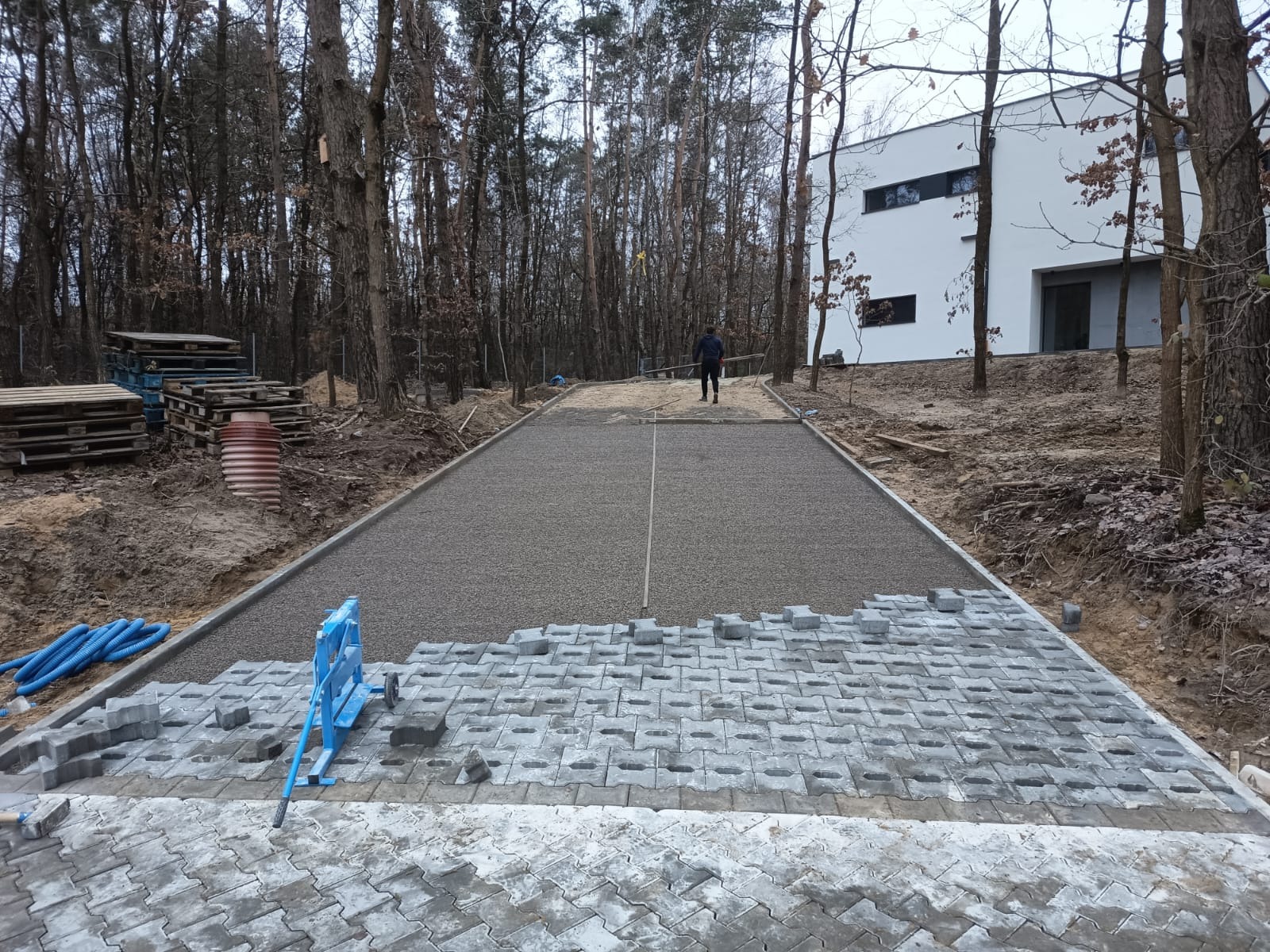 Układanie szarej kostki brukowej typu ażur na podjeździe do nowoczesnego domu w otoczeniu lasu. Widoczny etap prac z użyciem niebieskiego urządzenia do cięcia kostki.
