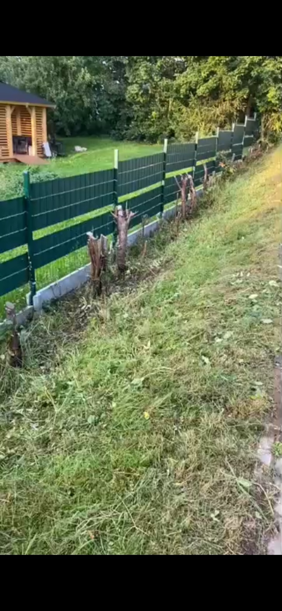 Nowoczesne, zielone ogrodzenie panelowe na betonowej podmurówce, widoczne pniaki po wyciętych krzewach, w tle altana ogrodowa i drzewa.