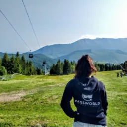 Osoba w bluzie z logo firmy na tle gór, kolejki linowej i zielonej łąki. Logo firmy Password Travels & Events.