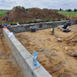 Fundamenty domu w trakcie budowy, z widocznymi bloczkami i butelką wody. W tle sterta ziemi i krajobraz wiejski pod pochmurnym niebem.