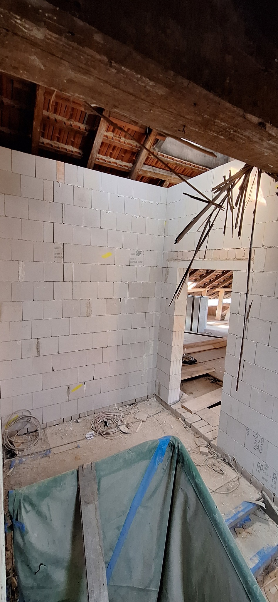 Wnętrze budynku w trakcie murowania ścian z bloczków. Widoczne fragmenty drewnianej konstrukcji dachu i instalacje elektryczne. Częściowo przykryte folią zabezpieczającą.