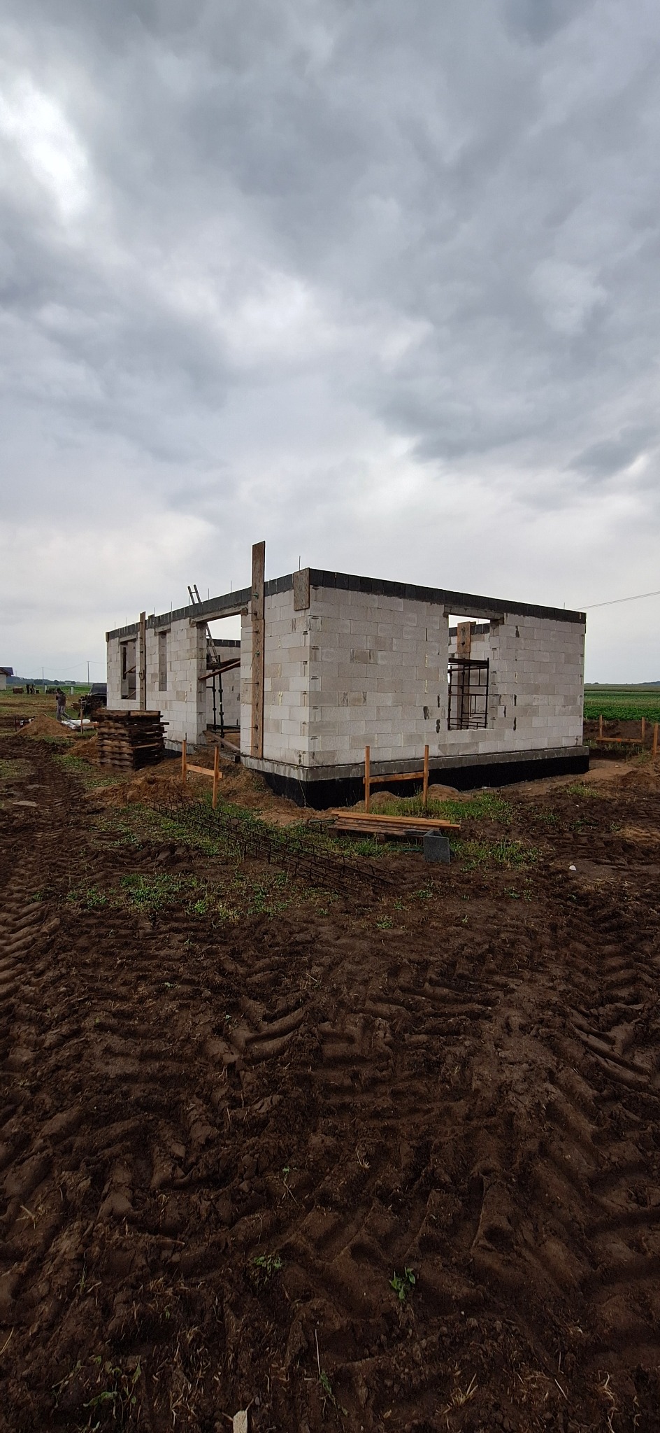 Niedokończony dom z betonu komórkowego pod pochmurnym niebem. Widoczne ślady opon na ziemi, drewniane podpory i drabina oparta o ścianę. Etap budowy domu.