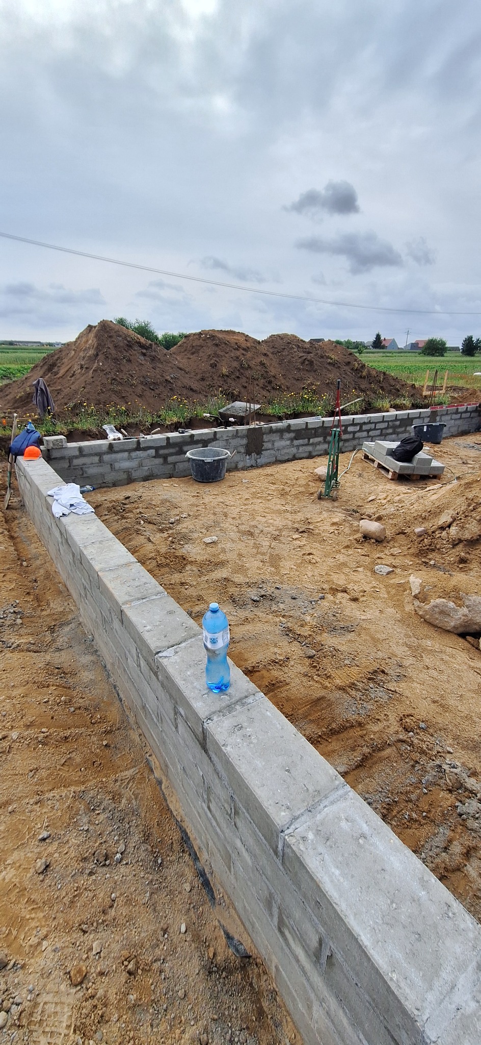Fundamenty domu w trakcie budowy, z widocznymi bloczkami i butelką wody. W tle sterta ziemi i krajobraz wiejski pod pochmurnym niebem.
