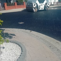 Star Budowa Inwestycje Sp z o.o. - Walec drogowy HAMM ubija świeżo wylany asfalt na ulicy, widoczne domy w tle. Obrzeże chodnika z kostki brukowej i białego kamienia.