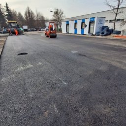 Star Budowa Inwestycje Sp z o.o. - Walec drogowy ubija świeżo wylaną, czarną nawierzchnię asfaltową na drodze. W tle budynek modułowy i pracownicy przy maszynie do asfaltu.