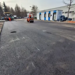 Walec drogowy ubija świeżo wylaną, czarną nawierzchnię asfaltową na drodze. W tle budynek modułowy i pracownicy przy maszynie do asfaltu.