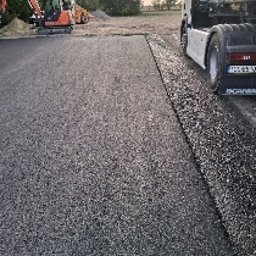 Star Budowa Inwestycje Sp z o.o. - Fragment nowo położonej, czarnej nawierzchni asfaltowej, z widocznym obrzeżem, ciężarówką i sprzętem budowlanym w tle, w słoneczny dzień.